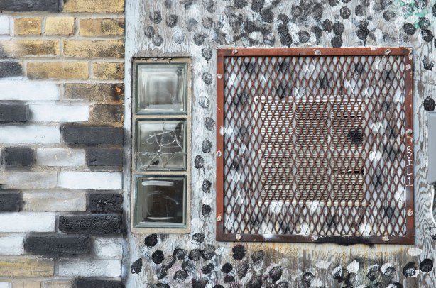 Three small windows arranged vertically beside a rusted metal grille covering an air vent.  Black and white splotches have been painted around the window. 