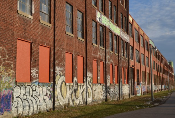 Three storey brick factory building. The windows in the lower storey are covered with orange wood. There is graffiti along the lower five or six feet of the building.