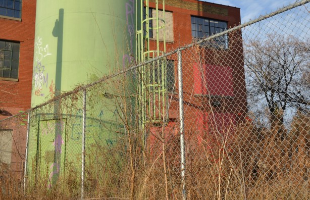a chainlink fence is in the foreground. Behind the fence is a tall green storage tank (the top part of it is not in the picture. One wall of a three storey red brick building is in the background.
