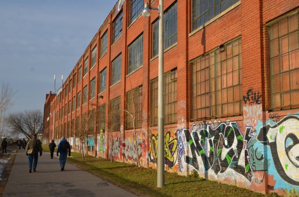 Looking north along the side of the old Viceroy rubber factory building. A red brick building with large windows. There is graffiti on the bottom five or six feet of the building.