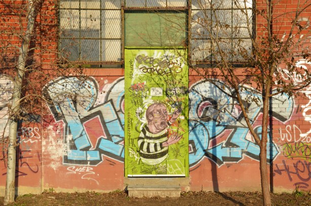 an old door is painted bright green and on it is a piece of street art of a man in a black and white striped shirt. A blue and white graffiti tag is on either side of the door.