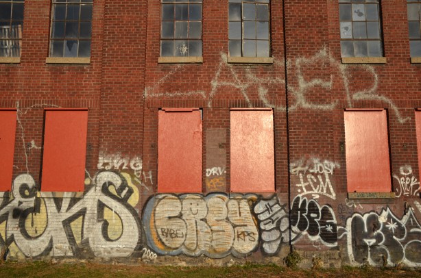 bottom two storeys of a red brick factory. The lower storey has the windows boarded up and painted orange. There are graffiti tags along the bottom of the building.