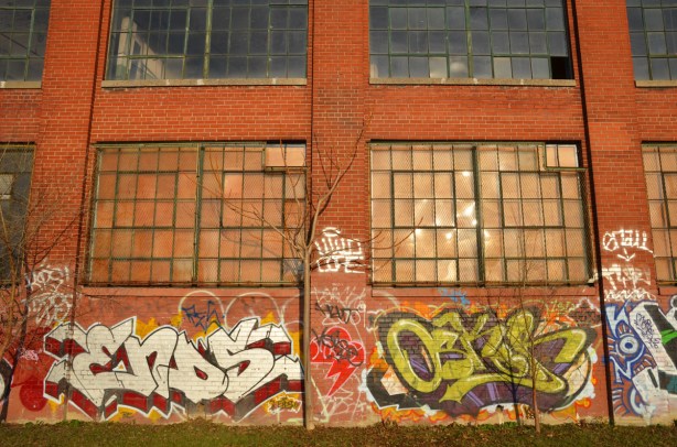The sun is shining on a red brick building with large windows. Graffiti tags are on the wall.