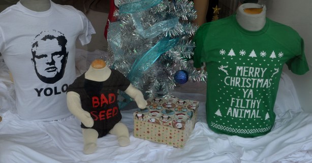 A window display in a T-shirt shop.  THere is a T-shirt with a picture of ROb FOrd with the word yolo printed on it.  There is a black toddler onsie with the words Bad Seed written in red.  There is a green T-shirt with the words Merry Christmas Ya Filthy Animal written on it. 