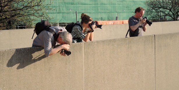 Three photographs are shooting from behind a low concrete wall.  One has his camera aimed downwards towards the people walking below.  The others have their cameras aimed farther out. 