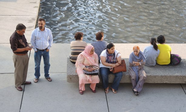 A group of people are either sitting on a bench, or standing beside it, in Nathan Phillips Square, beside the fountain pool 