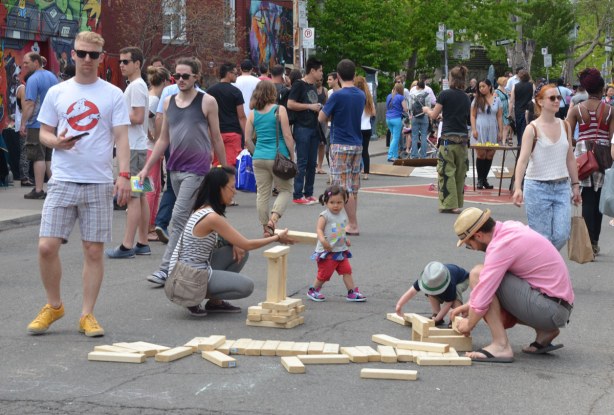 A pile of large wood blocks are in the middle of the street and a family is building small towers with them - a small girl and boy and their parents.