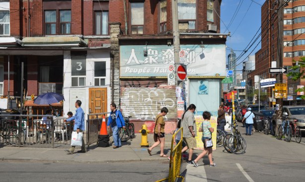 street scene of the intersection of Kensington and Dundas West. There are people walking on the sidewalk past a boarded up shop.