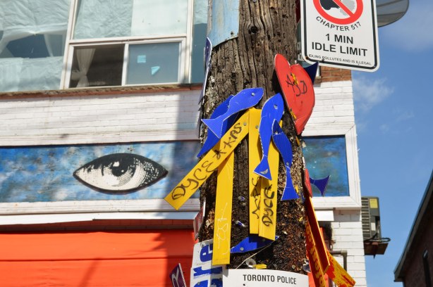 Detail of messages attached to a telephone pole. They are written on strips of yellow and blue heavy paper. In the background is a painting of a large eye, part of the sign above the door of Orbital Arts on Augusta Ave.
