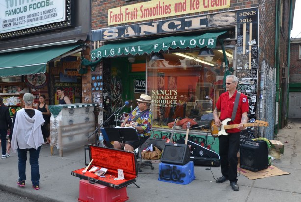 Two men on the sidewalk outside Sancis store. One man is standing and the other is sitting.