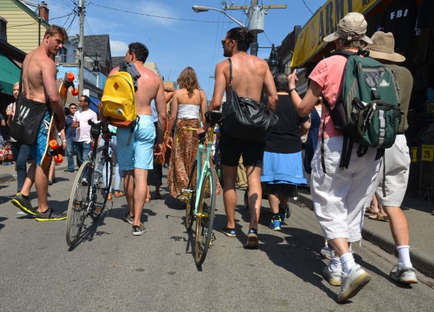 A group of young people are walking down the street, away from the camera. Some are walking their bikes and some of the men are topless.
