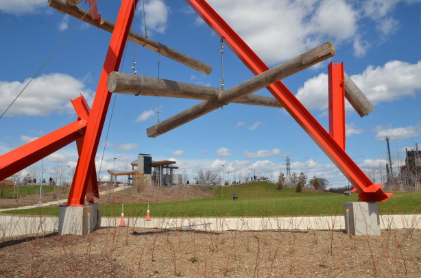 Part of a large red metal sculpture is in the foreground, looking past it you can see the pavillaion on top of a small hill that is part of Corktown Common park 