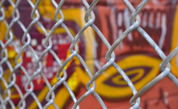 A chain link fence in front of a piece of graffiti in oranges, yellows and purple 