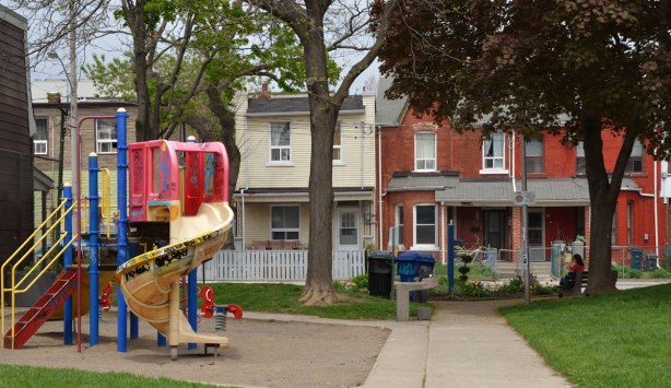 A play structure with slide is on the left of the picture, a woman is sitting on a bench on the right side.  Row houses on the opposite side of the street can also be seen.  There are some large trees too. 