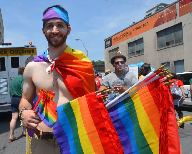 A man is wearing a rainbow flag like a cape, and he has one wrapped around his head like a headband.  He is selling large and small sized rainbow flags.  He is topless otherwise 
