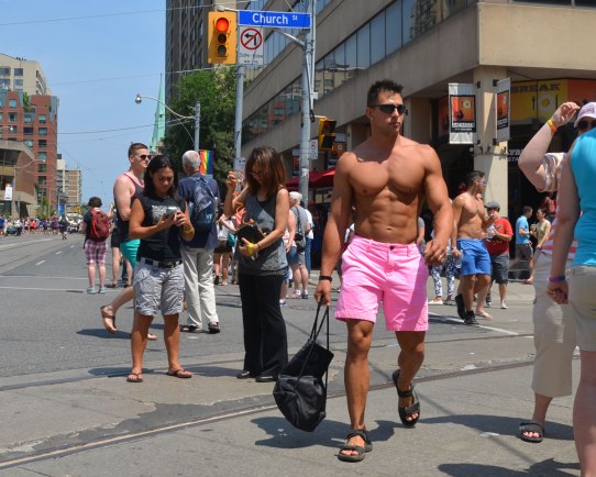 A good looking, very fit, man wearing just a pair of pink shorts and a pair of sandals is walking down the street. 