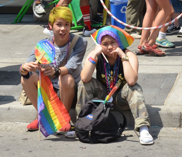 Two guys are sitting on the sidewalk.  One has dyed his hair yellow and he's carrying a rainbow flag.  The other is resting his chin on his hands, and his elbows on his knees.  He has his rainbow flag draped over his head and he looks very bored.  