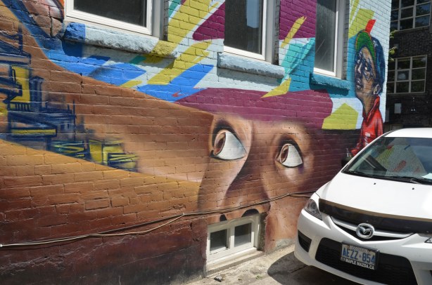 graffiti on a brick wall on Bulwer St., big white eyes on a brown face looking up towards the sky. A white Hyundai is parked beside it.