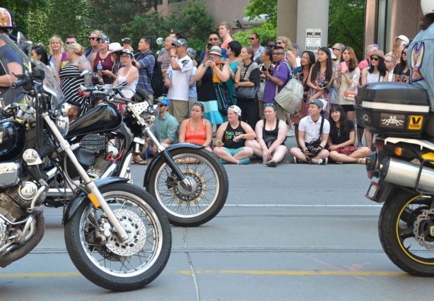 A crowd is watching the parade from the sidewalk.  Motorcycles are passing by