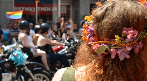 In the foreground of the photo is the back of a woman's head.  She has long wavy reddish hair and she is wearing a garland of flowers in her hair.  In the background are motorcycles in the parade. 