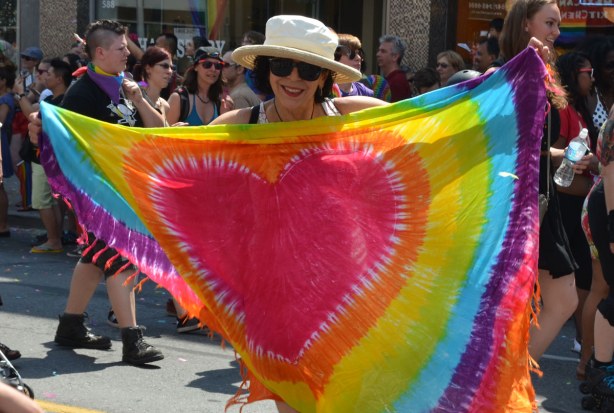 A happy looking woman holding up a large rainbow coloured scarf that has a bright red heart in the middle of it 