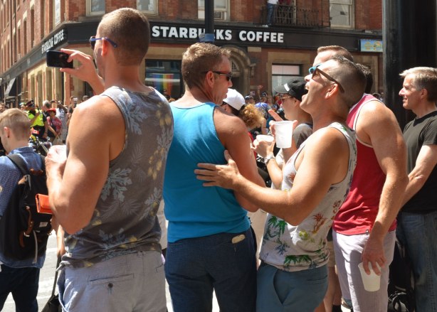 A group of men is standing beside the street, watching the parade.  One is using his phone to take a photo.  One is leaning his head back and laughing.