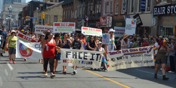 a group of women marching in the parade with a banner that says Face It, other walkers are holding up signs that say Stop Homophobia and Transphobia
