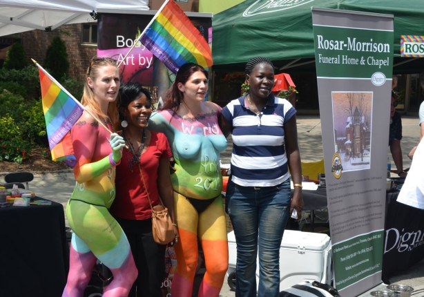Two topless women are painted in big rainbow colours. Two clothed women are posing for a photo with them.