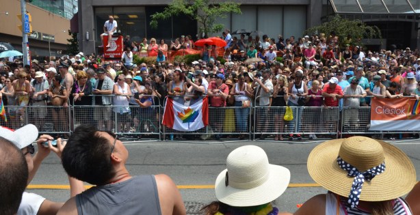 A row of people wearing hats in the foreground, looking across an empty street to a very large crowd behind barricades