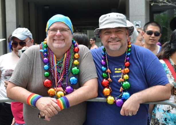 Two men are standing beind, and leaning on, the barricade. They are wearing multicoloured beads, incuding some very big beads that look more like balls for a Christmas tree