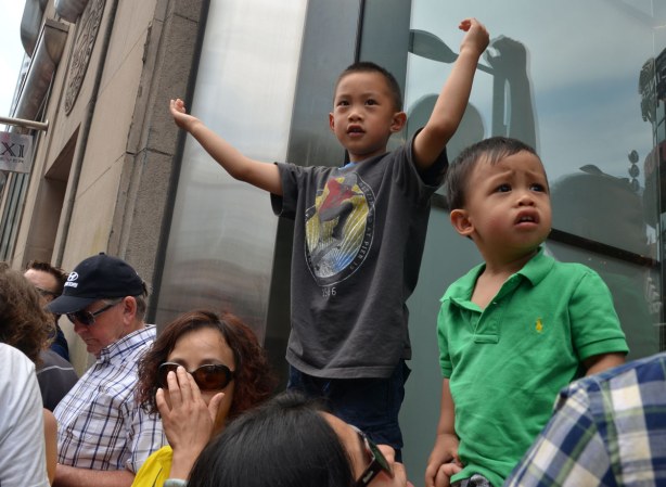 Two boys are standing on a window ledge so they are above a crowd of people. The small boy doesn't look too happy.
