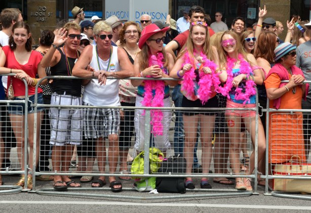 A group of people are standing behind metal barricades waiting for the parade to start, some are waving and some are smiling. A group of three girls are wearing pink boas.