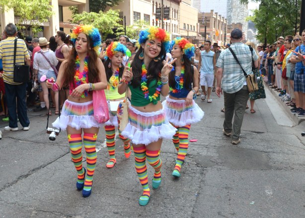 a group of four girls wearing rainbow curly wigs, frilly short skirts and rainbow striped socks are walking up the street between two rows of parade watchers