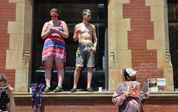 Two people are standing on the window sill of a large window.  The woman is wearing a striped dress.  She is texting.  The guy beside her is topless, wearing grey shorts