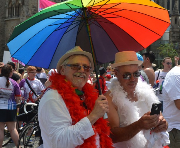 Two older men dressed in white are under a rainbow striped umbrella. One has a white boa, the other has a red boa.