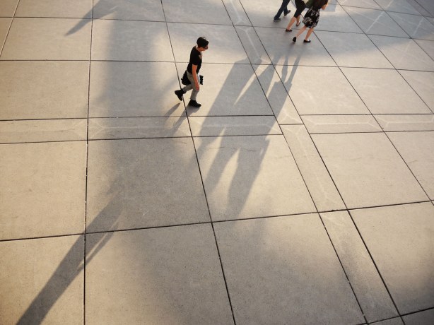 Most of the picture is of shadows on the concrete below.  There are partial shadows of the arches over the fountain at nathan phillips square.  There are also 4 people (or parts of the people) and the long shadows that they cast. 
