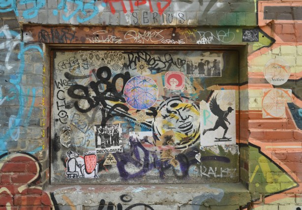 A window space in a brick wall that was covered over a long time ago.  Now there are tags and stickers covering the space as well as graffiti on the walls around the space. 