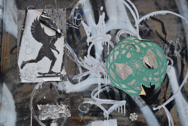 two stickers on a tag & scribble covered space.  One sticker is a silhouette of a man on a skateboard.  He has large wings coming out of his back.  Ths other sticker is a greenish colour geometric design within a greyish white circle. 