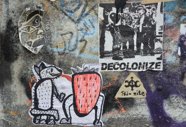 four stickers on a wall.  One is a mask that looks either African or BC First Nations.  One is a hairy dog like creature with a long tongue that is licking a red strawberry with legs.  One has the word 'decolonize' on it along with a picture of a group of men.  The last sticker is just the word 'phi-nite' with a symbol. 