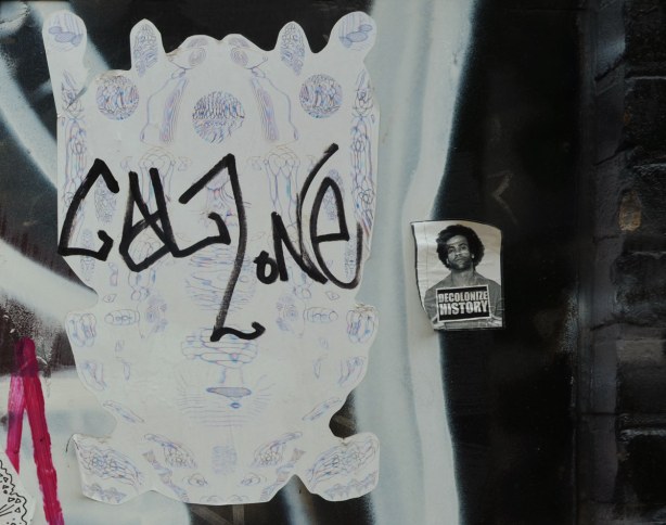 a purple and white sticker that has been defaced by the tag 'calzone'.  Beside it is a sticker that says 'decolonize history' with a mug shot of a man. 