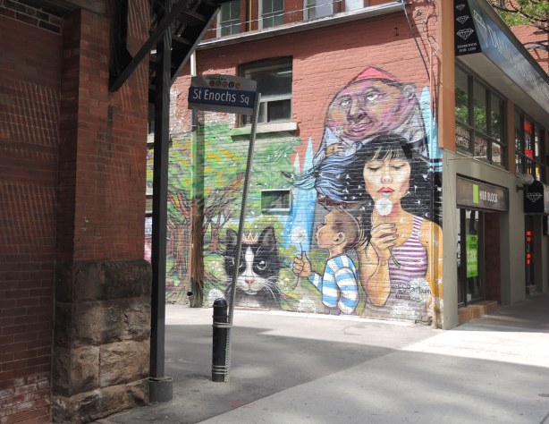 entrance to laneway, red brick building on the left (Massey Hall), mural painted on the wall on the right. Mural shows a woman & a boy blowing the seeds from white puffy dandelions.