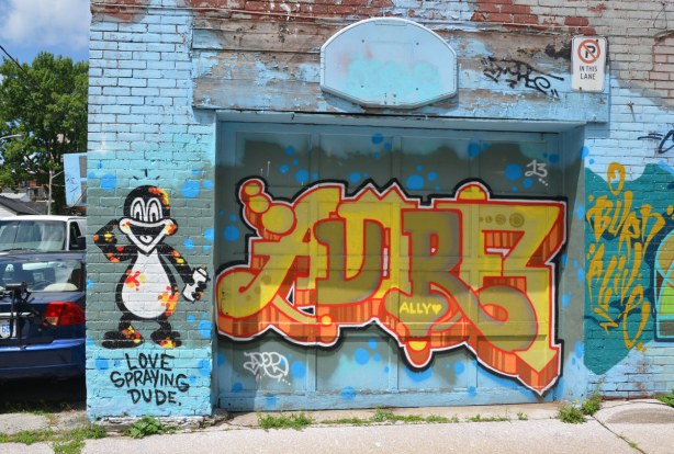Graffiti, or street art, on a blue background, of a little black and white animal with a spray paint can in his hand.  Beside him is a tag in yellows and olive greens. 