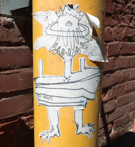 A black and white sticker on a yellow pole.  The words on the sticker say Rad Brad.  It is weird looking two legged creature. 