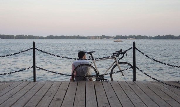 A cyclist and his bike are sitting at the edge of Lake Ontario, behind the chain fence.  It is evening and the sky is strating to turn pink 