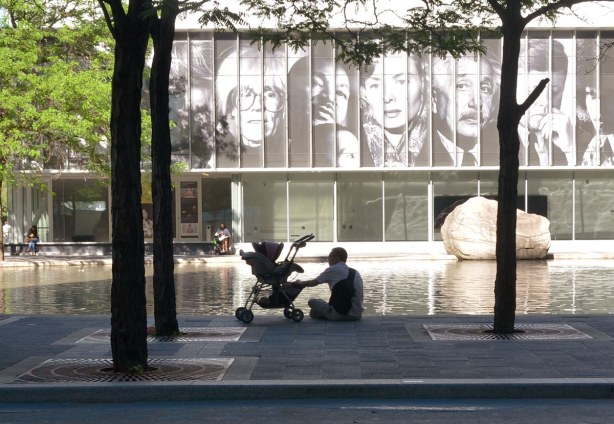 A man is sitting beside a stroller in the foreground.  Behind him is a pool of water and then a large glass building.   On the wall of the building are black and white images of some famous people including Andy Warhol, JFK, and Albert Einstein 