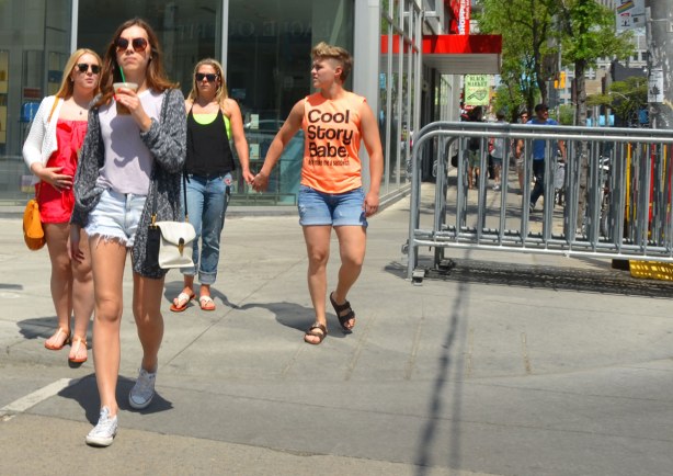 Some people are walking towards the camera as the cross a street including a couple who are holding hands.  He is wearing an orange Tshirt that says Cool story babe, Now Make me a Sandwich. 