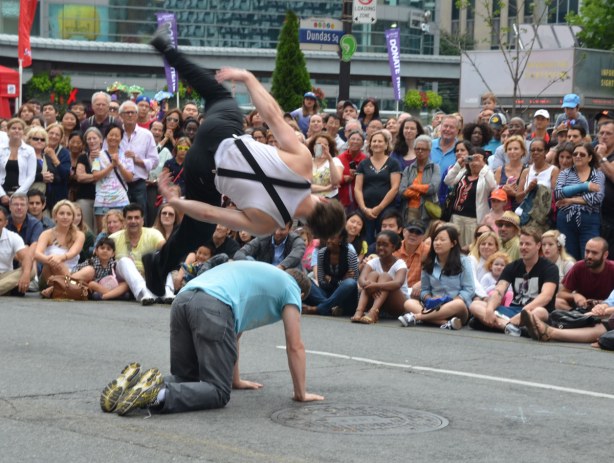 One of the street performers from the group Touch2Catch is doing a side flip over a man who is on all fours on the street.
