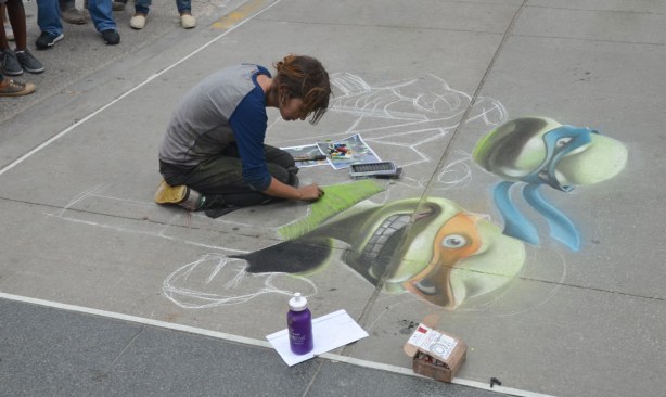 A woman is using chalk to make a large picture of the Teenage Mutant Ninja Turtles on the sidewalk
