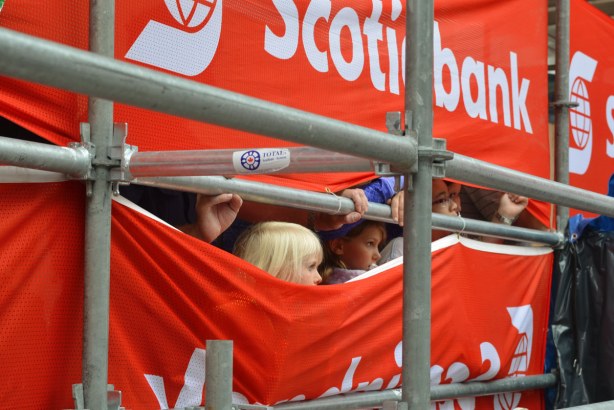 Some children are watching buskers perform by peeking through a gap in the red and white plastic banners that are the backdrop to the show.