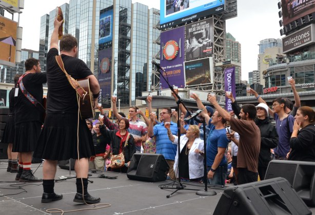 Musicians onstage, a group of adults standing in front of the stage. The adults all have drinks in their hands and they are all toasting. One of the musicians has a stuffed fish in his hand.